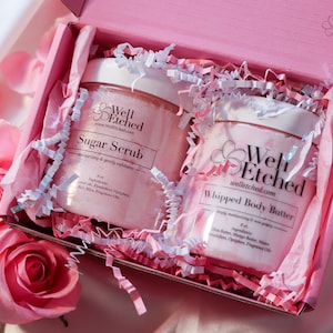 May include: Two pink jars of body care products, a sugar scrub and a whipped body butter, are nestled in a pink gift box with white tissue paper. The jars are labeled "Well Etched" and feature the brand's website address.