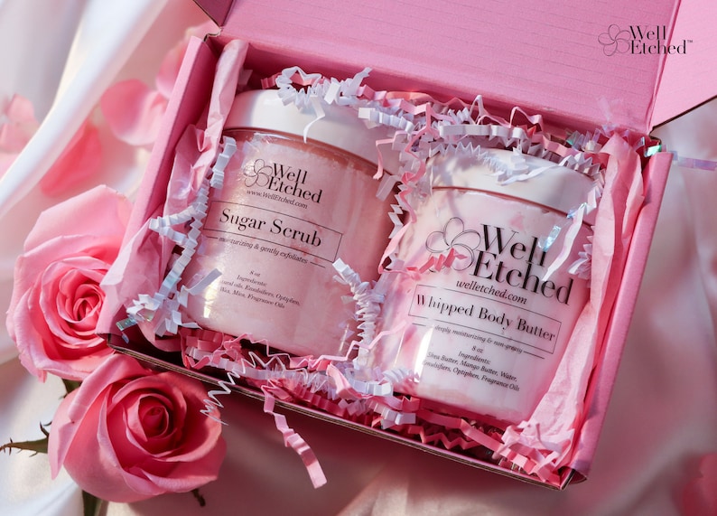May include: Two pink jars of body care products, a sugar scrub and a whipped body butter, are nestled in a pink gift box with white tissue paper. The jars are labeled "Well Etched" and feature the brand's website address.