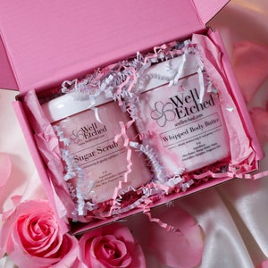 May include: A pink gift box with white tissue paper and two jars of body care products. The jars are labeled "Well Etched" and contain "Sugar Scrub" and "Whipped Body Butter".