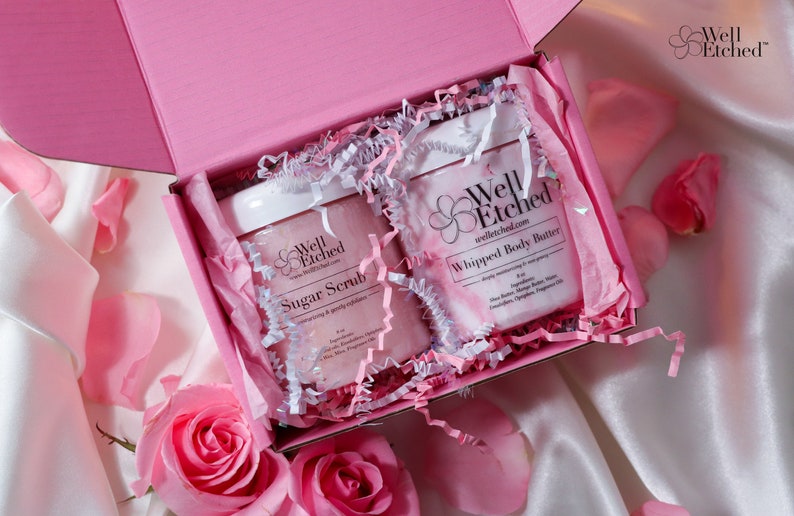 May include: A pink gift box with white tissue paper and two jars of body care products. The jars are labeled "Well Etched" and contain "Sugar Scrub" and "Whipped Body Butter".