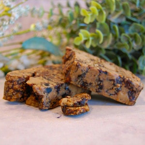 May include: Close-up of several slices of a baked good, possibly bread or cake, with a speckled appearance. The slices are a mix of light and dark brown, with visible seeds or inclusions. Green foliage is visible in the background.