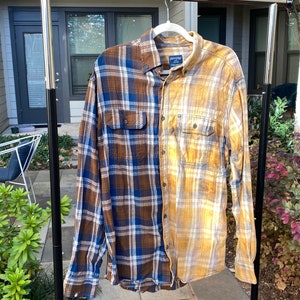 May include: A long-sleeved plaid shirt, half brown and blue, the other half yellow and tan, hangs on a white hanger. The shirt has two chest pockets and button closures. The shirt is displayed on a black metal clothing rack outdoors.