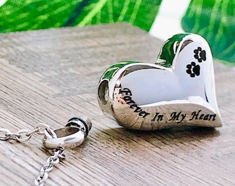 May include: A silver heart-shaped pendant with the words "Forever In My Heart" engraved on it. The pendant has three black paw prints and comes with a silver chain. The background is a wooden surface with green leaves.