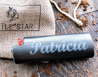 May include: A dark grey lighter engraved with the name "Patricia" in a cursive font. Beside it is a small burlap bag with the text "LITTLE STAR" and a unicorn logo. The background is a wooden surface.
