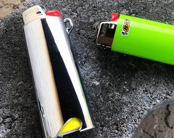 May include: Two BIC lighters on a textured surface. One is chrome with a red button, the other is bright green with the BIC logo. Both lighters have a red ignition button and a yellow fuel indicator.