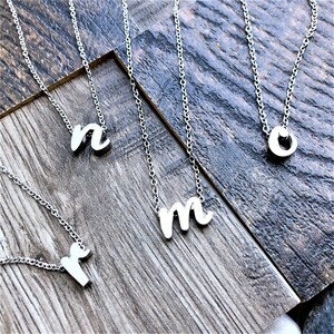 May include: Silver-toned initial necklaces with delicate chains. The letters 'n', 'm', 'o', and 'r' are displayed. The necklaces are arranged on a rustic, wooden surface, creating a simple and elegant aesthetic. These necklaces are perfect for personalized gifts.