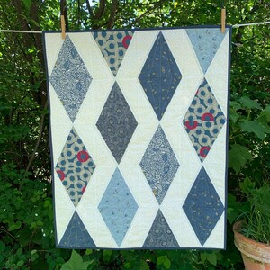 May include: A handmade quilt with a diamond pattern. The quilt features various blue and cream-coloured fabrics, with diamond shapes in different patterns and shades of blue. The quilt is hanging outside on a washing line, held by wooden pegs.