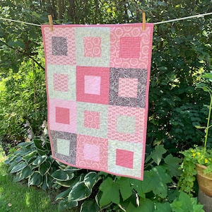 May include: A pink and grey patchwork quilt with a floral pattern. The quilt is hanging on a clothesline outdoors.
