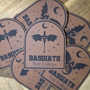 May include: Brown leather patches with a black dragon and crescent moon design. The patches have the text "BASGIATH War College" in a bold font. The patches are in an arched shape with a dragon and star design.