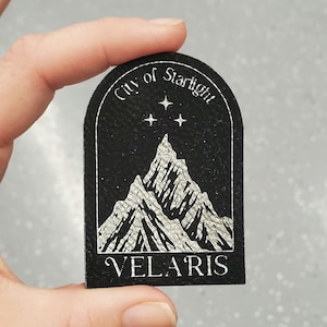 May include: Black patch with a white mountain range design, three stars, and the words "City of Starlight" and "VELARIS". The patch is held by a hand.