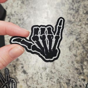 May include: Black and silver patch in the shape of a skeleton hand making the "hang loose" gesture. The patch has a black background with silver detailing on the bones. The patch is held up by a person's fingers.