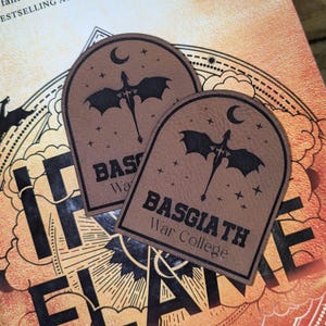May include: Two brown leather patches with a dragon and crescent moon design. The text "BASGIATH War College" is printed in black. The patches are on a book with the title "IF THE FLAME".