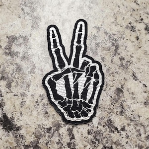 May include: A black and white patch in the shape of a hand making the peace sign. The hand is a skeleton hand with the bones visible. The patch has a black border and is on a textured background.