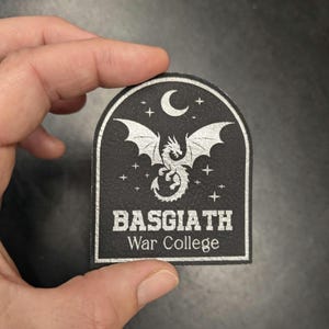 May include: Black patch with a silver dragon design, crescent moon, and stars. The text "BASGIATH War College" is printed below the dragon. The patch has a textured surface and a rounded top.