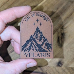 May include: A brown leather patch with the words "City of Starlight" arched across the top and "VELARIS" at the bottom. A black mountain range graphic is in the center, with three small stars above it. The patch is held by a hand.