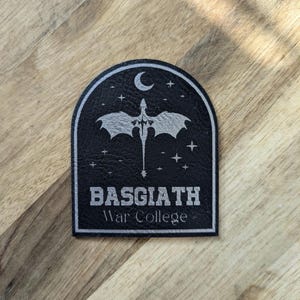 May include: Black patch with a silver design featuring a bat-like creature, stars, and a crescent moon. The text "BASGIATH War College" is displayed in silver lettering. The patch has a rectangular shape with rounded corners.