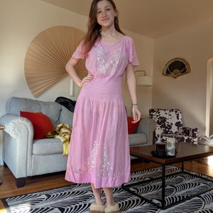 May include: A light pink dress with white floral embroidery. The dress features short sleeves, a fitted bodice, and a flowing skirt. The model is wearing brown slippers.