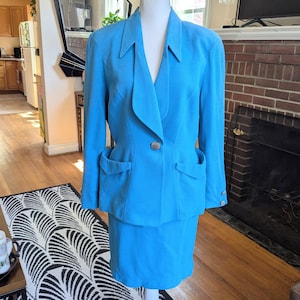May include: A bright turquoise skirt suit displayed on a white mannequin. The suit features a blazer with a notched collar, two front pockets, and a decorative button. The matching skirt is knee-length. The suit is made of a smooth fabric.