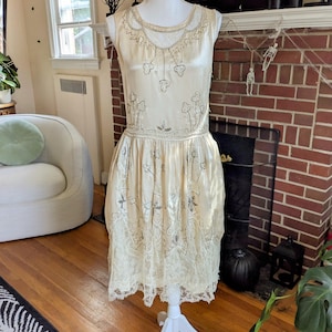 Puede incluir: Vestido vintage color crema con intrincados abalorios y ribete de encaje. El vestido sin mangas presenta un escote redondo y una cintura baja, con adornos de cuentas en forma de flores. El vestido se exhibe en un maniquí blanco.