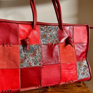 Puede incluir: Un bolso tote grande de cuero rojo con un diseño de patchwork. El bolso tiene varios compartimentos y está hecho de diferentes texturas de cuero, incluyendo gamuza. El bolso tiene un asa superior y se cierra con una cremallera.