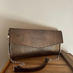 May include: A bronze-colored clutch with a textured surface and a fold-over flap. The bag features a gold chain strap and a matching brown leather handle. The clutch is resting on a wooden surface.
