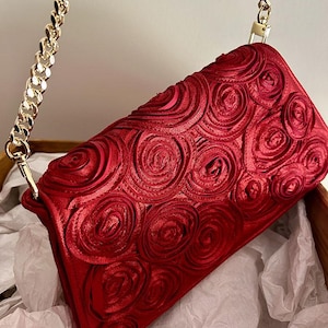 Puede incluir: Un bolso de mano de satén rojo con una correa de cadena dorada. El bolso está decorado con un patrón de rosas en 3D.