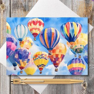 May include: Watercolor art print of colorful hot air balloons against a blue sky. The balloons are in various colors, including red, yellow, blue, and purple. The print is on a white card, with a gold pen resting below.