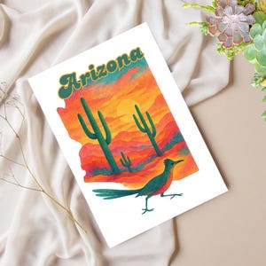 May include: A white print featuring the state of Arizona with a sunset landscape, cacti, and a roadrunner. The word "Arizona" is in green and gold lettering. The artwork is on a white background.
