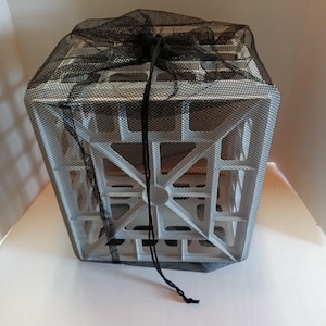 May include: A silver-coloured, square-shaped plastic crate with a geometric, open-grid structural design. The crate is enclosed within a black, fine-mesh drawstring bag that is tied at the top with thin black cords hanging down the front side.