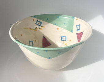 Może przedstawiać: Biała ceramiczna miska z geometrycznym wzorem w odcieniach niebieskiego, zielonego, czerwonego i żółtego. Miska ma szeroki rant i zaokrąglone dno.