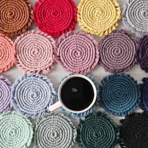 May include: A collection of colorful crocheted coasters with fringe, featuring various colors like pink, blue, green, yellow, and brown. A cup of coffee sits on top of one of the coasters.