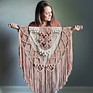 May include: A large macrame wall hanging with a geometric design in shades of pink and white. The hanging features a central diamond shape with fringe details.