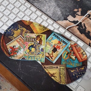May include: Sleep mask with a brown background and colorful book covers. The covers feature titles like "Pinocchio," "Fables," and "Peter Pan." The mask has a white elastic band and is resting on a keyboard.