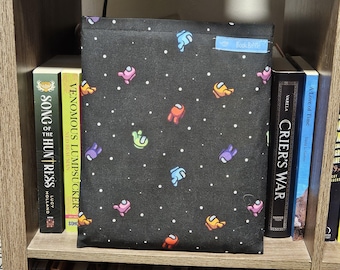May include: A black fabric storage box with a white polka dot pattern and colorful Among Us characters. The box is sitting on a bookshelf with other books.