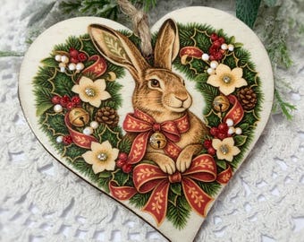 May include: A heart-shaped wooden ornament featuring a brown rabbit illustration centered within a wreath of green holly, red berries, white flowers, and pinecones. A red ribbon with a small gold bell is tied around the rabbit's neck.