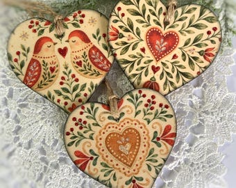 May include: Three heart-shaped ornaments with cream backgrounds, green foliage, and red accents. One features two birds, another a central red heart, and the third a decorative orange heart. Each ornament hangs from a brown twine loop.