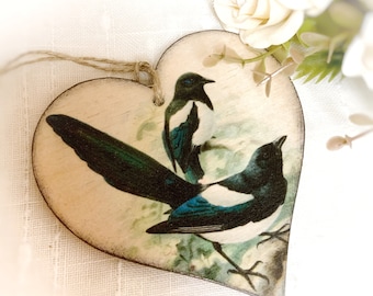 May include: Heart-shaped wooden ornament featuring a detailed illustration of two magpie birds. The birds have black and white plumage with teal accents. A jute string is attached for hanging. Decorative floral elements are in the background.