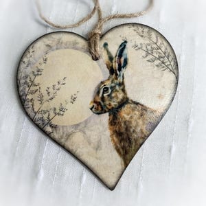 May include: Heart-shaped wooden ornament featuring a detailed illustration of a brown hare. The hare is set against a soft, neutral background with a circular design element. The ornament is adorned with a jute twine for hanging.