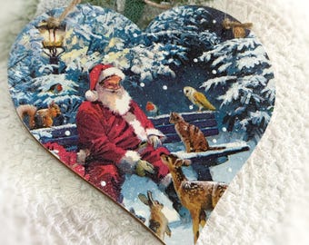 May include: Heart-shaped Christmas ornament featuring Santa Claus in a snowy forest scene. Santa is seated on a bench with a cat, deer, and other woodland creatures. The ornament has a string for hanging.