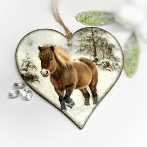 May include: Heart-shaped wooden ornament depicting a brown horse in a snowy scene. The horse has a light mane and tail. The ornament has a twine loop for hanging and is decorated with glitter.