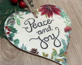 May include: Heart-shaped wooden Christmas ornament with the words "Peace and Joy" in black script. The ornament is decorated with a poinsettia, holly, and other greenery. A jute string is attached for hanging.