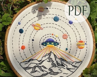 May include: Embroidered hoop art featuring a colorful solar system design with planets, dotted lines, and a UFO above a mountain range. The text "PDF" is visible. The artwork is set in a wooden hoop.
