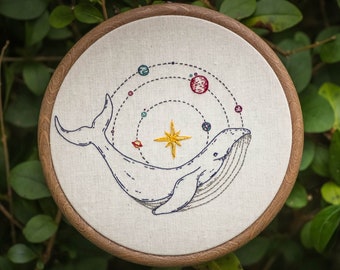 Könnte beinhalten: Stickbild mit einer gestickten weißen Wal, in dessen Körper ein stilisiertes Sonnensystem dargestellt ist. Der Wal ist mit schwarzem Faden umrandet und das Sonnensystem besteht aus bunten Planeten und einem gelben Stern.