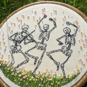 May include: Embroidered hoop art featuring three skeletons dancing in a field of flowers. The skeletons are black and white, and the flowers are a mix of orange, yellow, and white.
