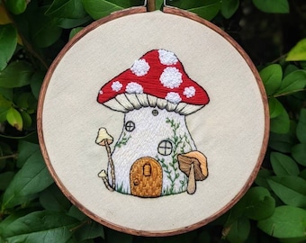 May include: Embroidered hoop art featuring a mushroom house with a red and white cap, a brown door, and green vines. The house is surrounded by small mushrooms.
