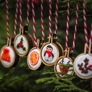 Może przedstawiać: Kolekcja ręcznie robionych ozdób choinkowych z różnymi wzorami haftowanymi, w tym pingwinem, cytrusami, ostrokrzewem i laską cukrową. Ozdoby są wykonane z drewna i mają rustykalny, vintage styl.