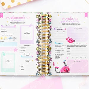 May include: Open planner with gold spiral binding, featuring colorful pages with text and illustrations. The pages are decorated with pink, blue, and yellow accents, with text in Spanish. The planner includes sections for planning and tracking goals.