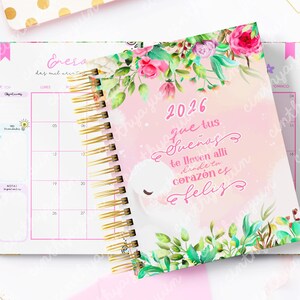 May include: A spiral-bound 2026 planner with a pink cover featuring floral accents and the text "2026 que tus sue&ntilde;os te lleven alli donde tu coraz&oacute;n es feliz". The open planner reveals a calendar layout with gold spiral binding.
