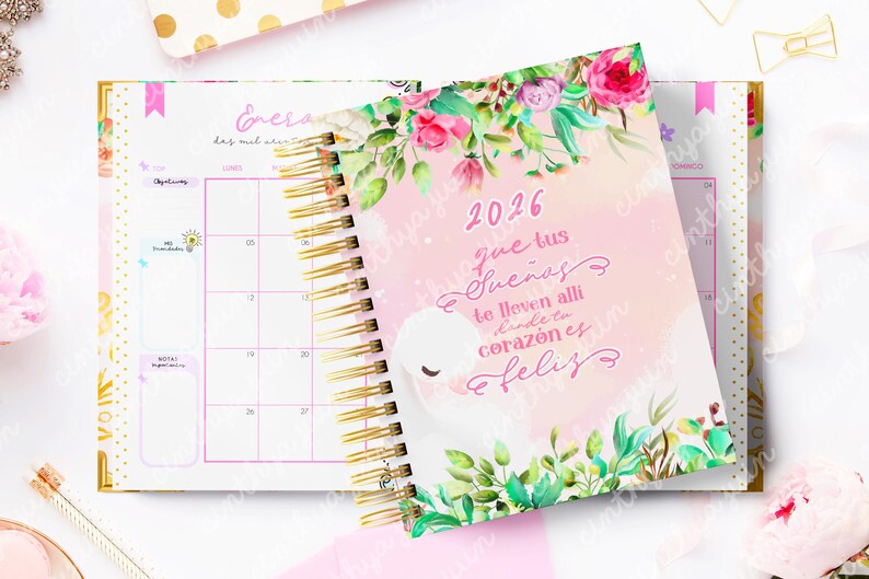 May include: A spiral-bound 2026 planner with a pink cover featuring floral accents and the text "2026 que tus sue&ntilde;os te lleven alli donde tu coraz&oacute;n es feliz". The open planner reveals a calendar layout with gold spiral binding.