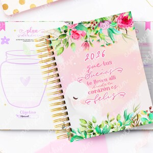 May include: A spiral-bound planner with a floral design and the year 2026 on the cover. The cover features a watercolor illustration with text in Spanish. The open planner shows lined pages and a jar illustration.